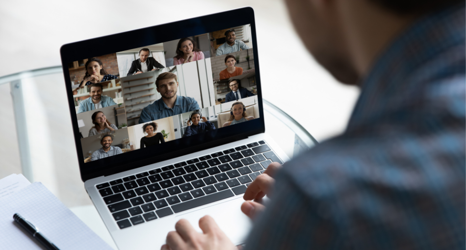 Over the shoulder photo of a bearded person on a video conference call with several other people. The other meeting invitees are all showing their faces in their individual environments on the call.
