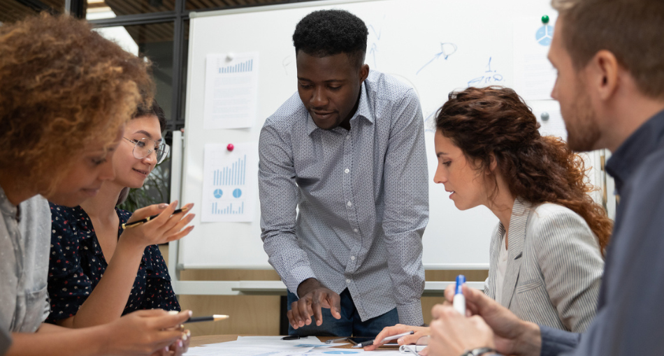 A man dressed in business casual is standing at the head of the table over some paperwork he's showing to his colleagues who are seated at the table. The colleagues are listening intently as plans are shared.