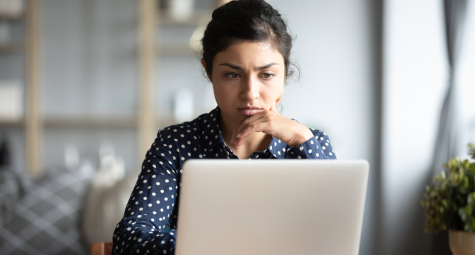 A woman frowns contemplatively while she reads something on the laptop screen in front of her.