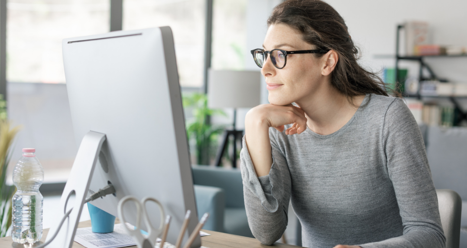 Photo of a woman sitting at desktop computer looking at the screen facing her, appearing confident about what she sees.