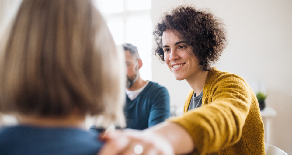 A woman smiles encouragingly to the person she's looking at, having placed her hand on this person's shoulder as a sign of support. We only see the back of this person in need of support.