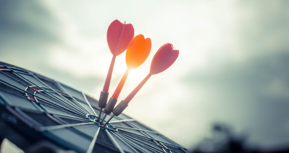 Three colorful darts are centered on the target of a dart board in front of a cloudy sky with the sun peeking through. Using predictive analytics while defining your ideal customer profile can give your team the clarity they need to act and drive business forward for new acquisition, retention, and expansion efforts.