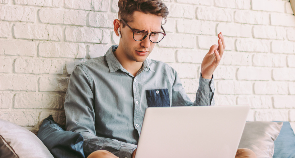 Man in glasses looking at his laptop in quizzical frustration, holding one hand up in question.