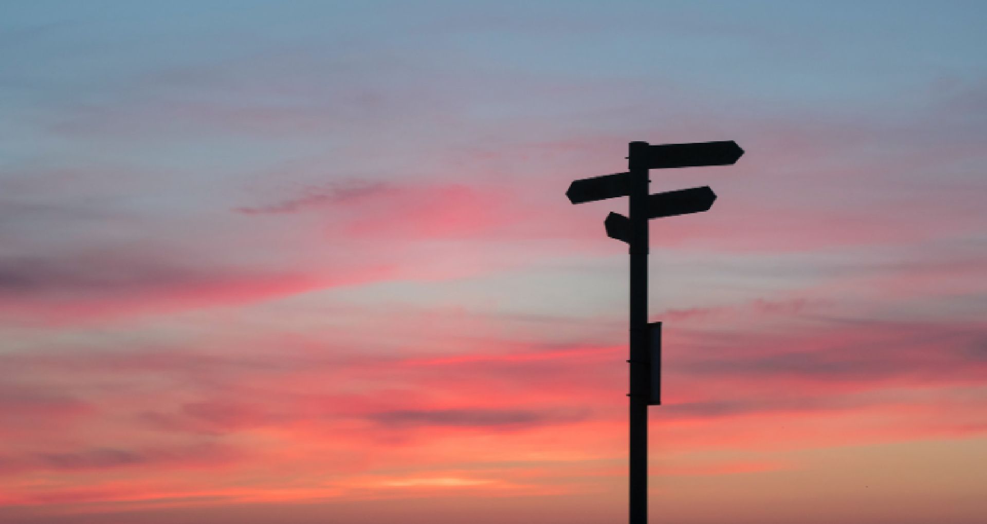 Sign post in the shadow with arrows pointing in different directions in front of a sunset sky.