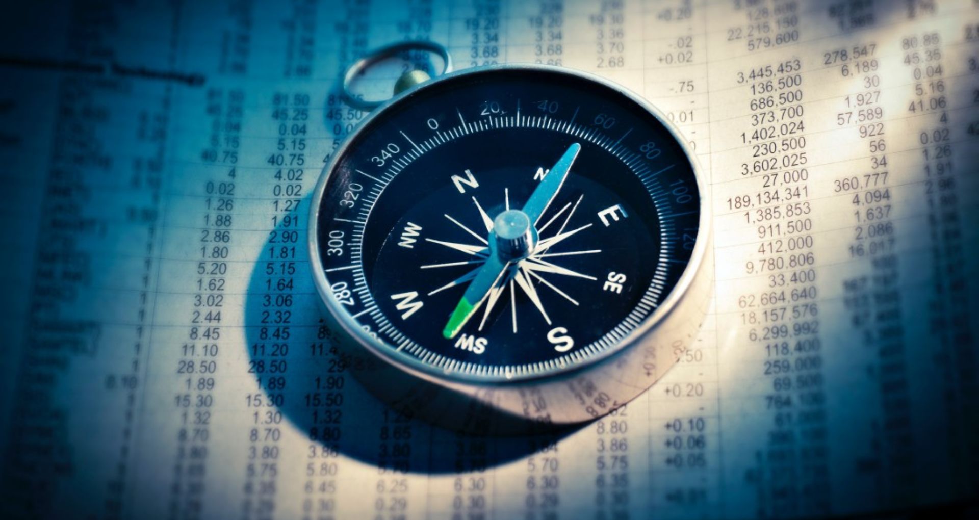 Picture of a compass under spotlight on top of a paper with lots of numbers in rows and columns.